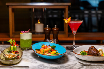 Elegant restaurant table with gourmet dishes, colorful cocktails on marble surface, ambient candlelight. Fine dining experience, sophisticated atmosphere, culinary artistry.