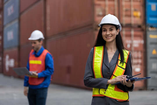 Smiling female logistic manager with hard hat and safety vest holding radio in cargo port with colleague in background