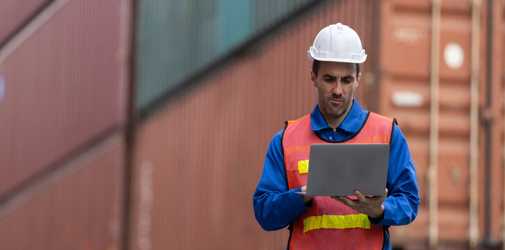 Focused young male worker wearing hard hat and safety vest uses laptop with concentration near shipping containers