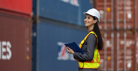 Smiling female engineer in hard hat and safety vest inspects cargo containers with confidence, overseeing logistics operations at shipping yard