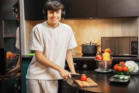 Happy teenage boy wearing headphones preparing a fresh vegetable salad at the kitchen table. Cutting tomatoes and vegetables, enjoying a healthy and cheerful cooking moment at home.