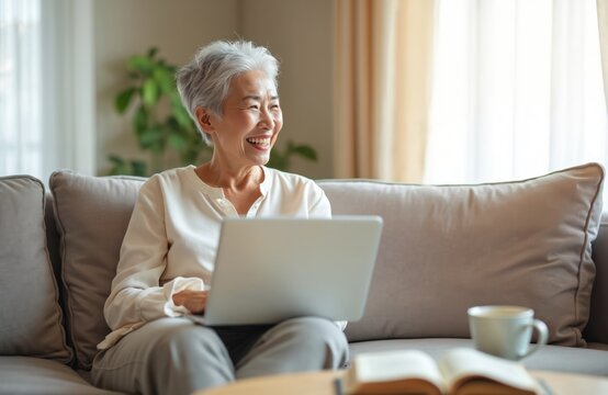 Cheerful senior Asian woman uses laptop on couch. Mature lady enjoys social media and technology. Smiling pensioner relaxes at home with computer in living room. Happy retirement lifestyle concept.