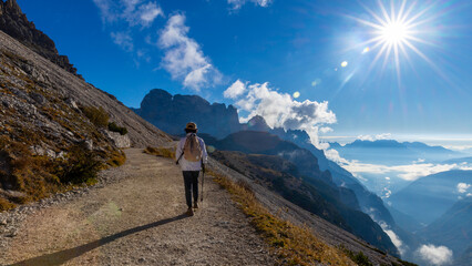 A senior traveler hikes alone along a mountain trail under bright sunlight, surrounded by stunning...