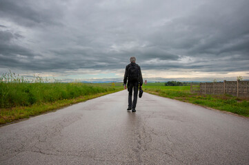 silhouette di uomo che cammina, in viaggio su strada, ripresa dal basso, che si perde all'infinito tra erba alta verso il cielo denso di nuvole.