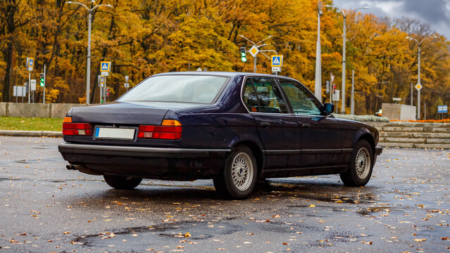 Blue classic sedan parked on a rainy autumn lot, set against wet trees with yellow foliage and covered with water droplets that create a calm, nostalgic atmosphere