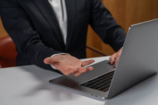 Businessman using laptop while gesturing with open hand during conversation in office environment, illustrating modern communication concept.