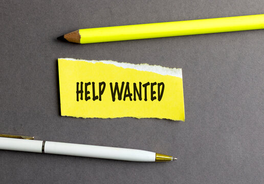 Help wanted message on torn yellow paper with pencil and pen on dark gray background