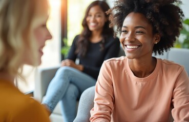 Three women sit, talk in bright space. Black lady smiles, engaging companion in conversation. Another person watches, also happy. Diverse group shares friendly moment, showing support, community.