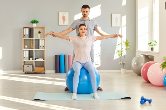 Male caregiver or health care worker helping woman patient doing sport exercises sitting on fit ball in rehabilitation center. Man physiotherapist helping girl in rehab. Physiotherapy concept.
