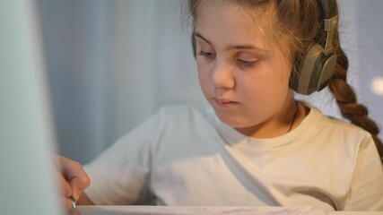 Girl in headphones writes in notebook. Kid focused on study with laptop. Student completes education tasks at desk. Learning supported by books and devices. Child continues focused education effort - Powered by Adobe