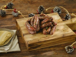 Homemade sausages are arranged on a wooden board, showcasing their rich texture and spices. A plate of mustard sits nearby, with pinecones and festive lights in the background