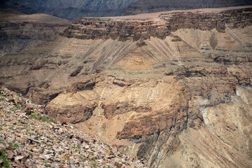 A dramatic view of the Fish River Canyon in Namibia, showcasing steep rocky cliffs, layered geological formations, and a winding riverbed cutting through the arid desert landscape. 