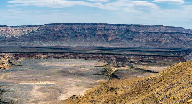 A dramatic view of the Fish River Canyon in Namibia, showcasing steep rocky cliffs, layered geological formations, and a winding riverbed cutting through the arid desert landscape. 