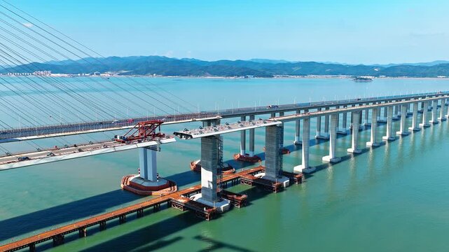 Aerial shot of a massive sea-crossing highway bridge under construction with engineering equipment over the water near a coastal city in China. 
