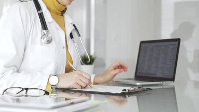 Doctor working on medical records, writing prescription in clinic office, using laptop for patient data management. Medicine, healthcare and science concept