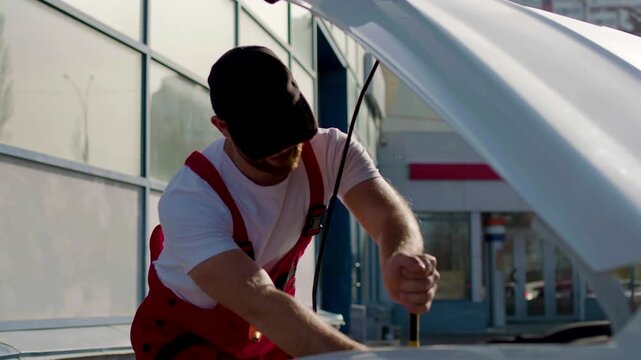 Auto mechanic in red overalls performing under-the-hood check on vehicle, inspecting engine components and fluid levels, showcasing automotive maintenance skills and expertise