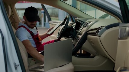 Mechanic in red overalls using a tablet inside a modern car, analyzing vehicle systems, showcasing advanced automotive technology and professional expertise