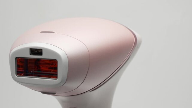 A modern pink IPL hair removal handset rotates slowly on a white background. Close side angle frames the flash window and smooth matte surface.