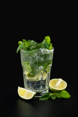 A tall glass filled with a sparkling beverage features fresh mint leaves and lime slices. Ice cubes keep the drink cool, creating a vibrant, refreshing look against a dark backdrop