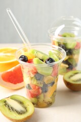 Tasty fruit salad in plastic cups and ingredients on beige table, closeup. Healthy snack