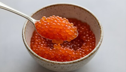 A close-up of vibrant red salmon caviar on a silver spoon over a ceramic bowl filled with more delicious roe