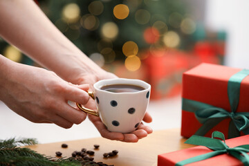 Woman holding cup of aromatic coffee above table with gift boxes, beans and fir tree branches...