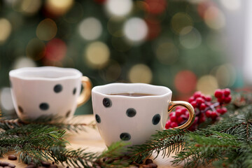 Aromatic coffee in cups, beans and fir tree branches on wooden table against blurred lights, closeup. Christmas greeting card