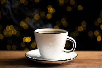 Aromatic coffee in white cup on wooden table against black background with blurred lights, closeup. Bokeh effect