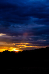 Obraz premium Dramatic golden sunset behind mountain silhouettes under deep moody blue clouds