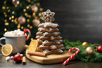 Christmas tree of gingerbread cookies, hot cocoa, cinnamon, dried orange slices and candy cane on table against dark background with blurred lights