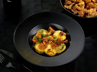 Ravioli filled with creamy cheese rests gracefully in a sleek black bowl, topped with savory sautéed mushrooms and fresh herbs, showcasing a gourmet meal