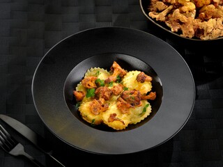 Delicious ravioli filled with cheese is elegantly served, topped with sautéed mushrooms and fresh herbs. The dark plate and contrasting colors create an inviting atmosphere for a delightful meal