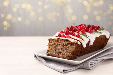 Tasty Christmas cake with icing, cranberries and rosemary on white wooden table against background with blurred lights, closeup