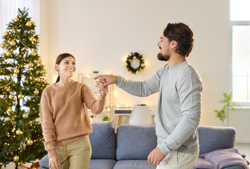 Christmas couple dancing at home, love moment of happy man and woman. Husband and wife dance to festive and joyful music, twirl by decorated Xmas tree in cozy living room, people sharing laughter