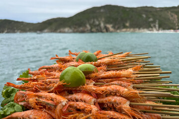 Close-up of grilled shrimp skewers topped with fresh lime halves, photographed against a blurred ocean background 