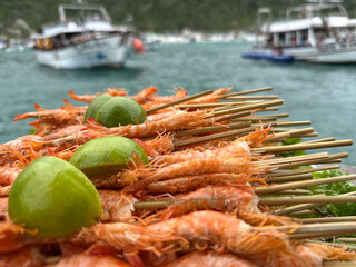 Close-up of grilled shrimp skewers topped with fresh lime halves, photographed against a blurred seaside background with boats on turquoise water.