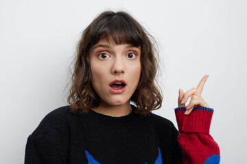 A surprised young woman stands against a light background, eyes wide, mouth open, and her index finger raised as if making a sudden discovery.