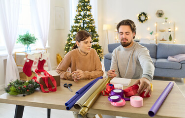 Happy couple wrapping gifts in boxes and foil with ribbons to prepare for Christmas celebrations at home. Man and woman siting at table in living room to wrap presents with Santa decor and paper