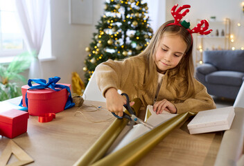 Child wrapping christmas gift near tree in living room. Little girl cuts paper at home with festive lights, preparing presents for family and santa during winter holiday season. Joyful celebration