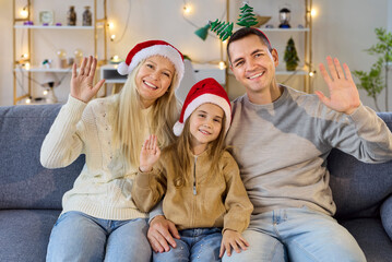 Christmas family video call, child waving happily. Parents and daughter in Santa hats sit on sofa at home, smiling at camera during online chatting. Internet connection on winter holiday.