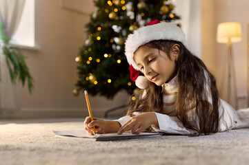Fototapeta premium Christmas child writing letter to Santa at home. Girl lies on floor by festive tree in santa hat, focused on wishlist with pen. Winter holiday and New Year celebration and preparations concept.
