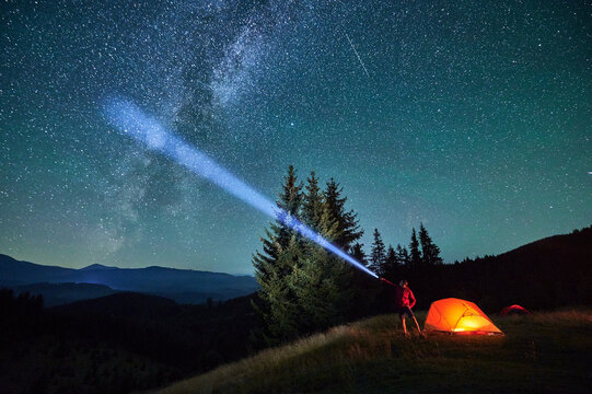 Man hiker stands under dazzling starry sky, shining flashlight into heavens. Milky Way stretches above, glowing tent nearby, creating magical and serene scene in peaceful mountain wilderness.