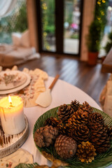 A festive table with a white tablecloth. On it are a plate with pine cones and candles, as well as other kitchen utensils.
