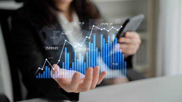 A businesswoman reviews financial data trends using a smartphone and digital graphs, showcasing modern technology in a dynamic office environment. Scalp