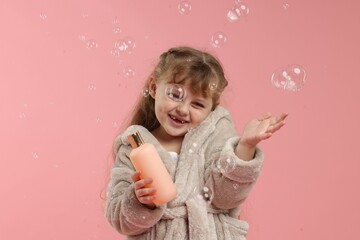 Cute girl in bathrobe with cosmetic product and bubbles on pink background