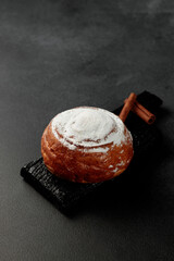 Round, delicious bun with cinnamon and powdered sugar on a black wooden board on a gray background