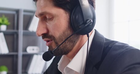 Closeup portrait of young business man working in call center at online support service office. Male operator agent in headset communicating with clients working in help desk agency. 4k video. - Powered by Adobe