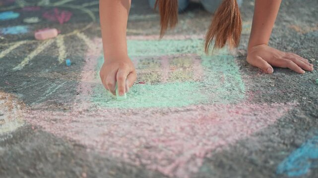 A girl kneels on the street to draw a house with chalk. Her hand shapes the house with steady lines, the chalk marking the asphalt as outdoor art and summer play fill the peaceful moment with joy.