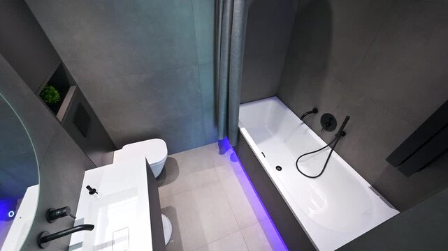 Modern bathroom with grey tiles, white fixtures, and minimalist design. Includes a round mirror with blue accent lighting, white sink, toilet, and bathtub with black hardware