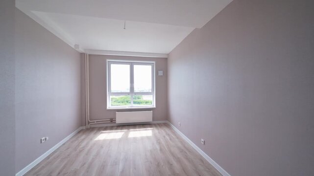 A bright, empty room with wooden floors and gray walls has a large window letting in sunlight.